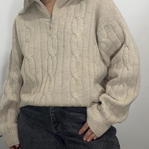 May include: A beige cable-knit sweater featuring a quarter-zip collar with a silver-toned metal zipper. The garment has long sleeves with ribbed cuffs and a vertical cable pattern throughout the front and sleeves, paired with dark gray denim jeans.