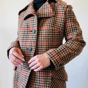 May include: A houndstooth patterned coat in brown, black, and red, featuring a notched collar and three black buttons. The coat has a classic design and is made of a woven fabric. The coat is a vintage style.