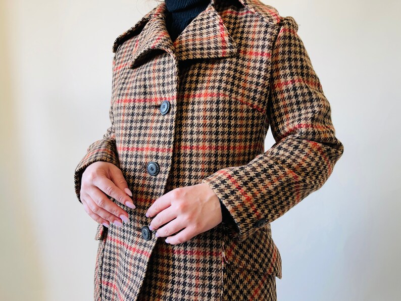 May include: A houndstooth patterned coat in brown, black, and red, featuring a notched collar and three black buttons. The coat has a classic design and is made of a woven fabric. The coat is a vintage style.