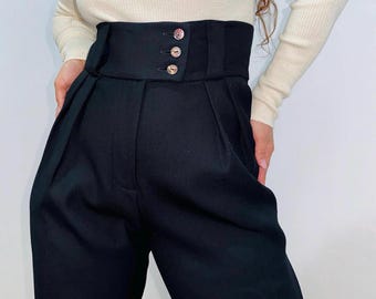 May include: High-waisted black trousers featuring a wide waistband with three circular buttons arranged vertically on the right side. The trousers have a structured fit with visible pleats extending from the waistband down the front legs.