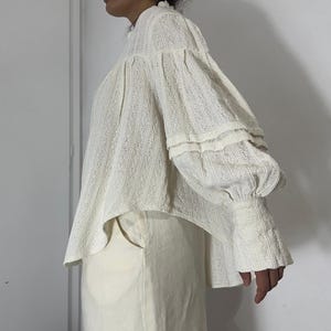 Puede incluir: Una blusa de manga larga en color blanco roto con tejido texturizado de rayas verticales. La prenda presenta un cuello alto, mangas abullonadas voluminosas con detalles fruncidos y pu&ntilde;os anchos y ajustados. El dobladillo es ligeramente curvo.