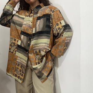 Peut inclure: Une chemise à manches longues boutonnée avec un motif patchwork dans les tons marron, noir et vert. La chemise a un col et des boutons dorés. Le tissu présente des motifs géométriques et floraux.