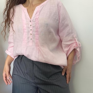 May include: A light pink linen blouse with a V-neck and button details. The sleeves are rolled up. The blouse is paired with gray pinstripe pants. The overall style is casual and comfortable.