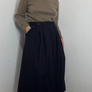 May include: A beige long-sleeve turtleneck sweater paired with a dark navy blue midi skirt. The skirt features a high waistband with a small metallic button detail and deep vertical pleats that create a full, flowing silhouette with side pockets.