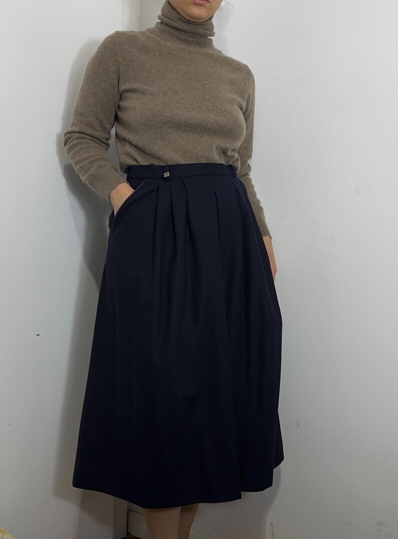 May include: A beige long-sleeve turtleneck sweater paired with a dark navy blue midi skirt. The skirt features a high waistband with a small metallic button detail and deep vertical pleats that create a full, flowing silhouette with side pockets.