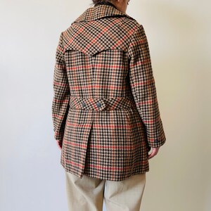 May include: A brown, black, and red check coat with a notched collar and a double-breasted design. The coat features a belt detail at the waist and a pleated back. The coat is worn over beige trousers.