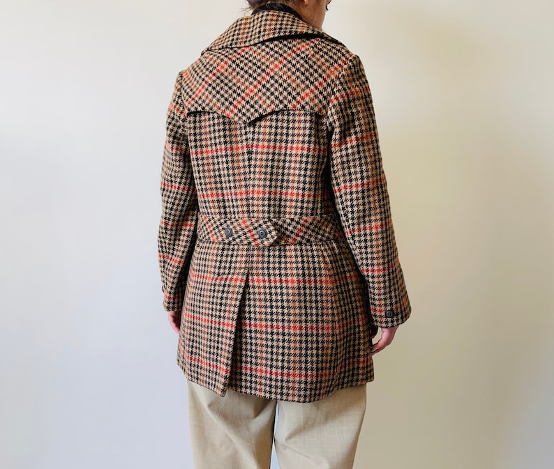 May include: A brown, black, and red check coat with a notched collar and a double-breasted design. The coat features a belt detail at the waist and a pleated back. The coat is worn over beige trousers.