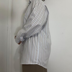 May include: A long-sleeved, button-down shirt with vertical stripes in light blue, white, and brown. The shirt has a chest pocket and is worn with a brown skirt. The fabric appears to be a lightweight material.