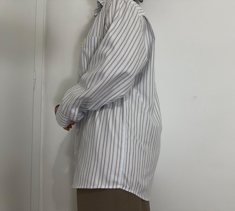 May include: A long-sleeved, button-down shirt with vertical stripes in light blue, white, and brown. The shirt has a chest pocket and is worn with a brown skirt. The fabric appears to be a lightweight material.