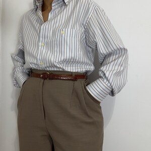 May include: A light blue and white striped button-down shirt with a chest pocket. The shirt is paired with brown trousers and a brown belt. The outfit is a classic, casual style.