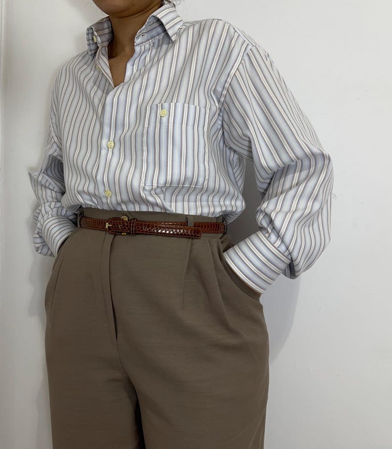 May include: A light blue and white striped button-down shirt with a chest pocket. The shirt is paired with brown trousers and a brown belt. The outfit is a classic, casual style.