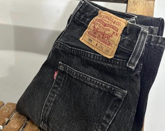 May include: Black denim jeans with a classic five-pocket design. The jeans feature a faded wash and a leather patch with the Levi Strauss & Co. logo. The size is 28W 30L. The jeans are folded on a wooden chair.
