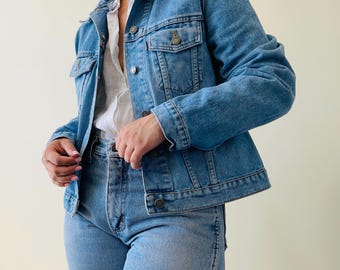 May include: Light blue denim jacket with button closures and two chest pockets. The jacket is paired with matching light blue denim jeans. The outfit is a classic, casual style.