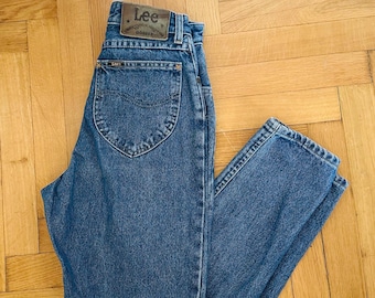 May include: Blue denim jeans with a classic five-pocket design. The jeans feature a leather patch with the "Lee" logo and the word "Gossip" on the back. The jeans are on a wooden surface.