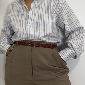 May include: A button-down shirt with vertical stripes in light blue, white, and gray, paired with brown trousers and a brown belt. The belt has a gold buckle. The shirt has a pocket on the chest.