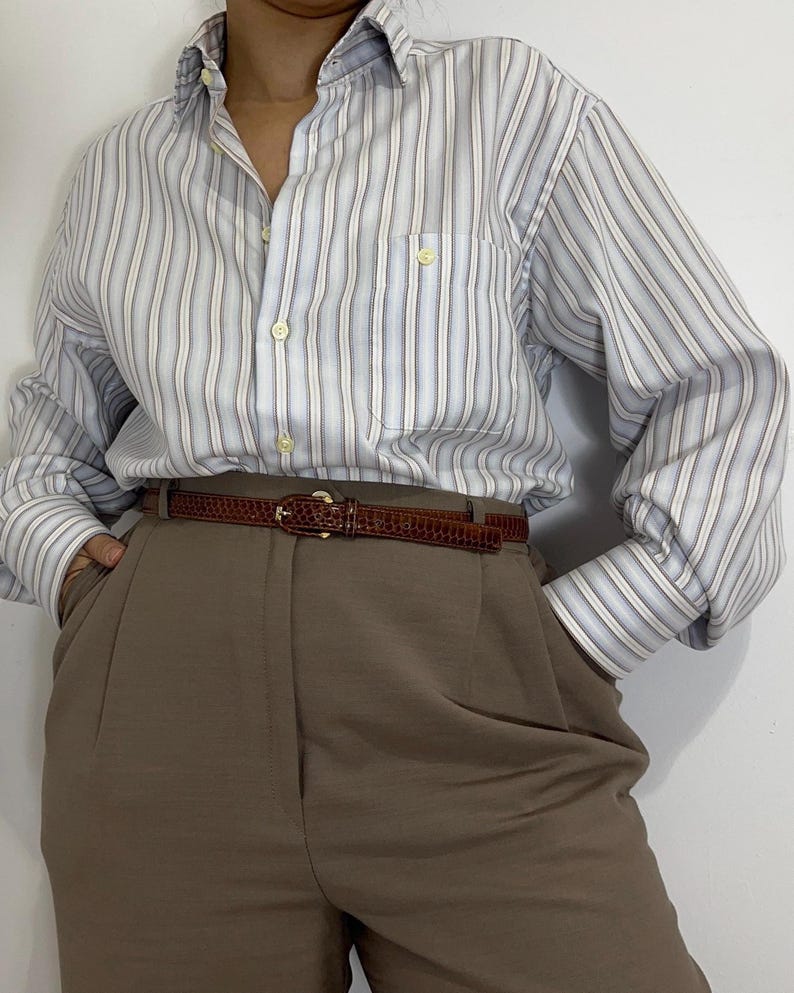 May include: A button-down shirt with vertical stripes in light blue, white, and gray, paired with brown trousers and a brown belt. The belt has a gold buckle. The shirt has a pocket on the chest.