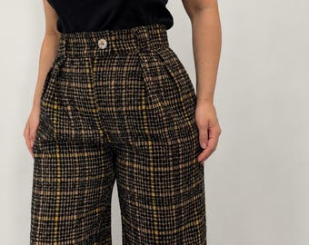 May include: High-waisted trousers featuring a black, tan, and yellow plaid pattern. The pants have a structured waistband with a single centered button, belt loops, and subtle pleats at the front. The fabric has a textured appearance.