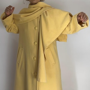 May include: A pale yellow coat with a matching scarf draped over the shoulders. The coat has a button-down front, two front pockets, and long sleeves. The coat is made of a soft, textured fabric.