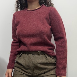 May include: A burgundy-colored sweater with a crew neck and long sleeves. The sweater has a soft, textured appearance. The model is wearing brown tweed pants with a button closure.