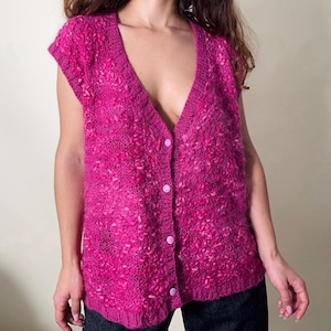 Può includere: Un gilet senza maniche in maglia fucsia con scollo a V e chiusura con bottoni. Il gilet ha un aspetto strutturato e nodoso ed è realizzato in un materiale morbido e soffice. I bottoni sono di colore fucsia abbinato.