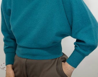 May include: A teal turtleneck sweater featuring long sleeves with dropped shoulders, a fitted ribbed waistband, and ribbed cuffs. The sweater is paired with solid brown trousers that have a visible front pocket and a structured waistband design.