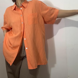 May include: An orange button-down shirt with short sleeves and a chest pocket. The shirt is made of a lightweight fabric and has a relaxed fit. The shirt is unbuttoned, revealing a glimpse of the wearer's pants.