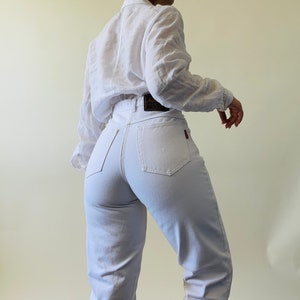 May include: A person wearing a white button-down shirt and light-colored jeans. The jeans have a high waist and a red logo on the back pocket. The shirt has long sleeves.
