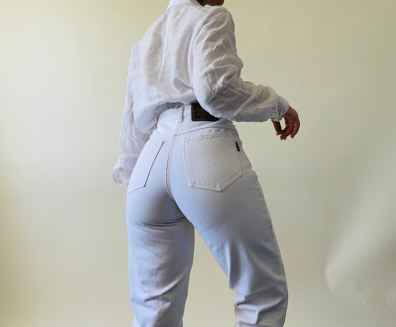 May include: A person wearing a white button-down shirt and light-colored jeans. The jeans have a high waist and a red logo on the back pocket. The shirt has long sleeves.