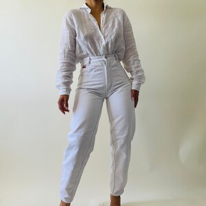 May include: A white button-down shirt and high-waisted white jeans. The shirt has long sleeves and a collar. The jeans have a straight leg fit and a red logo on the back pocket. The outfit is simple and classic.