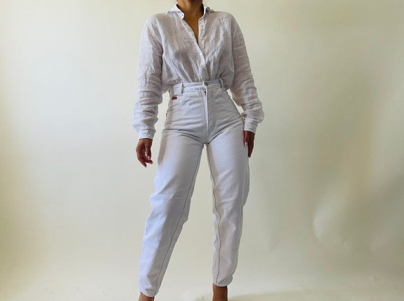 May include: A white button-down shirt and high-waisted white jeans. The shirt has long sleeves and a collar. The jeans have a straight leg fit and a red logo on the back pocket. The outfit is simple and classic.