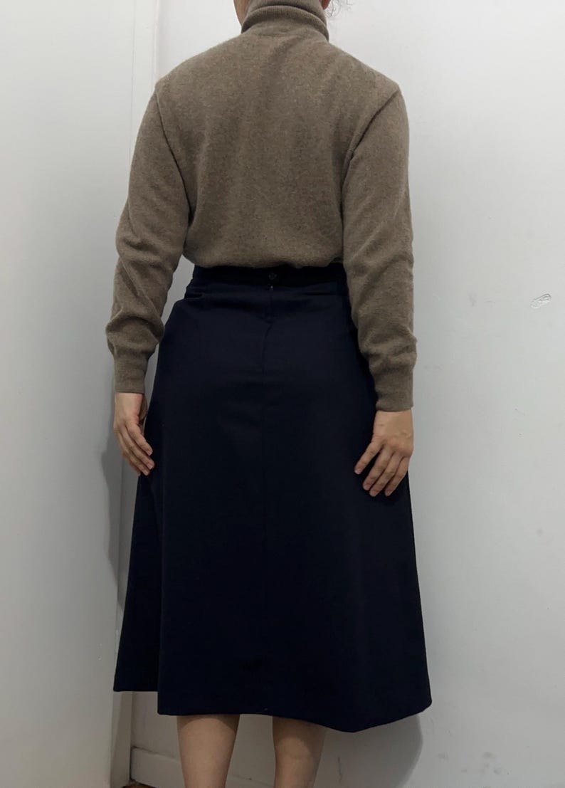May include: A beige long-sleeved turtleneck sweater paired with a solid navy blue midi-length skirt. The sweater features a ribbed collar and cuffs, while the skirt has a smooth, plain finish with a visible center seam and a button closure at the waist.