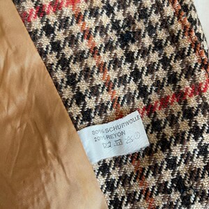 May include: Close-up of a houndstooth fabric in brown, black, and red. The fabric is part of a garment, with a tan lining visible. A white tag displays the text "80% SCHURWOLLE 20% RAYON".