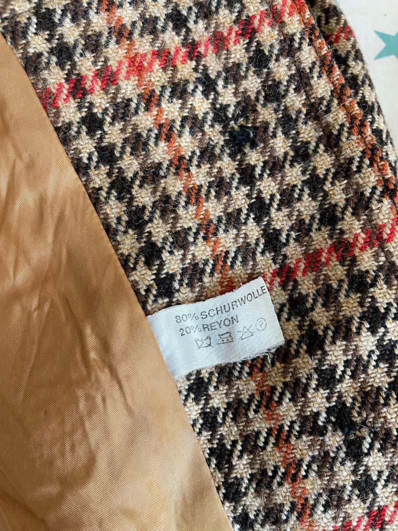 May include: Close-up of a houndstooth fabric in brown, black, and red. The fabric is part of a garment, with a tan lining visible. A white tag displays the text "80% SCHURWOLLE 20% RAYON".