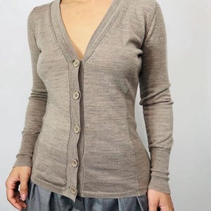 Könnte beinhalten: Hellbraune Strickjacke mit V-Ausschnitt und langen Ärmeln. Die Strickjacke hat eine Knopfleiste mit fünf Knöpfen. Der Stoff scheint ein feines Strick zu sein. Die Strickjacke wird mit einer grauen Hose getragen.