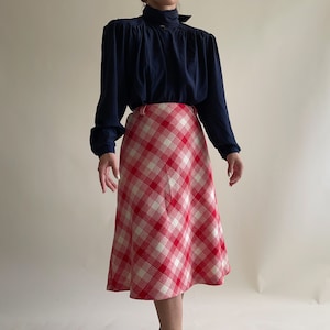 Pode incluir: Uma blusa azul marinho de manga comprida com gola alta e detalhes de botões é combinada com uma saia xadrez vermelha e branca. A saia, que vai até a panturrilha, cria um conjunto clássico de inspiração vintage. A saia tem cerca de 76 cm de comprimento.