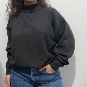 Könnte beinhalten: Ein dunkelgrauer Langarmpullover mit hohem Stehkragen und gerippten Bündchen. Das Strickkleidungsstück hat eine lockere Passform mit überschnittenen Schultern und wird mit einer blauen Jeans kombiniert.