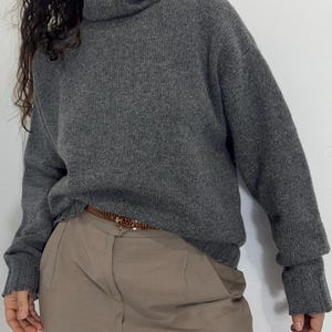 Könnte beinhalten: Ein grauer Rollkragenpullover mit gerippten Bündchen und Saum. Der Pullover wird mit einer beigefarbenen Hose und einem braunen Gürtel mit goldener Schnalle getragen. Das Outfit ist lässig und bequem.