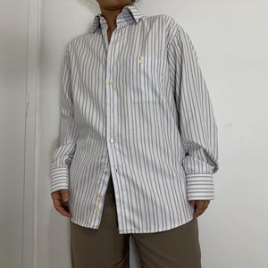 May include: A button-down shirt with vertical stripes in white, light blue, and brown. The shirt has a chest pocket and buttoned cuffs. The sleeves are rolled up. The shirt is paired with brown pants.