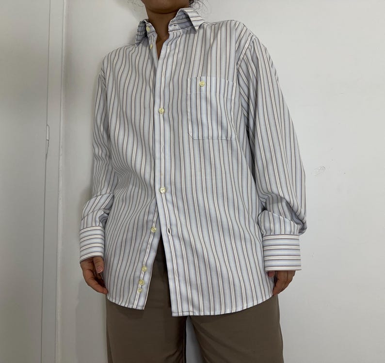 May include: A button-down shirt with vertical stripes in white, light blue, and brown. The shirt has a chest pocket and buttoned cuffs. The sleeves are rolled up. The shirt is paired with brown pants.