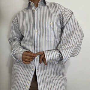 May include: A button-down shirt with vertical stripes in shades of white, light blue, and gray. The shirt has a chest pocket and buttoned cuffs. The collar is open, and the buttons are a light yellow color. The shirt is made of a lightweight fabric.