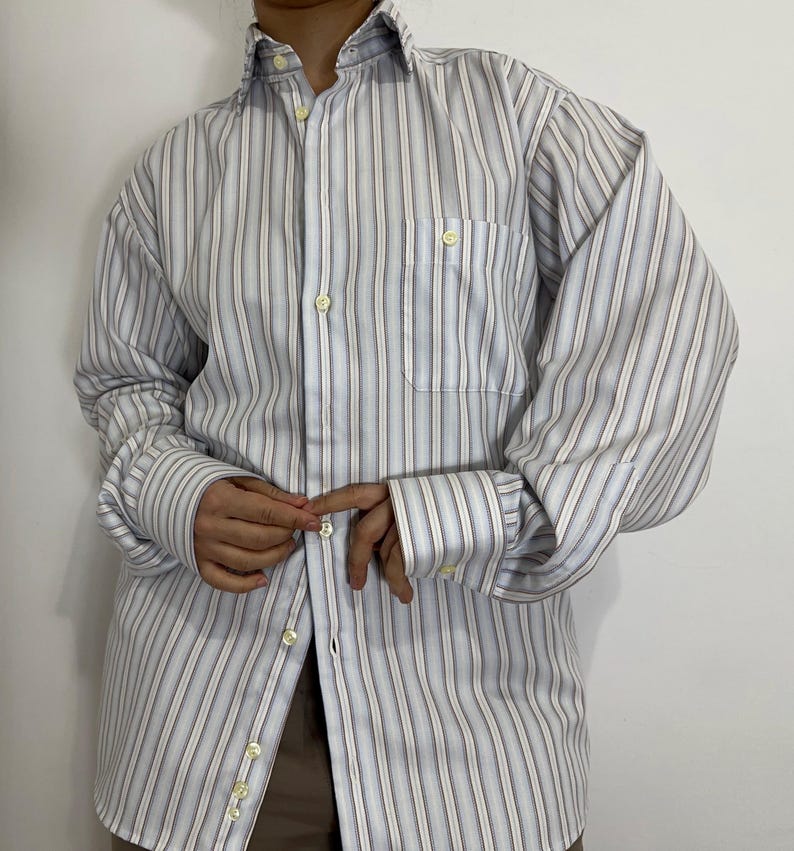 May include: A button-down shirt with vertical stripes in shades of white, light blue, and gray. The shirt has a chest pocket and buttoned cuffs. The collar is open, and the buttons are a light yellow color. The shirt is made of a lightweight fabric.