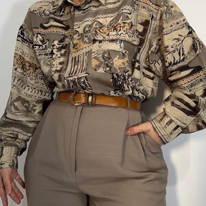 Könnte beinhalten: Ein langärmeliges Hemd mit Knopfleiste und einem architektonischen Druck in Beige, Braun und Creme, inklusive Brusttasche, kombiniert mit einer taubengrauen Hose mit hoher Taille und einem beigen Ledergürtel mit goldener Schnalle.