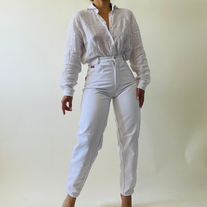May include: A white button-down shirt and high-waisted white jeans. The shirt has long sleeves and a collar. The jeans have a high waist and a straight leg. The outfit is made of a lightweight fabric.