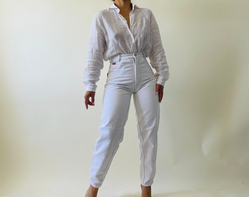 May include: A white button-down shirt and high-waisted white jeans. The shirt has long sleeves and a collar. The jeans have a high waist and a straight leg. The outfit is made of a lightweight fabric.