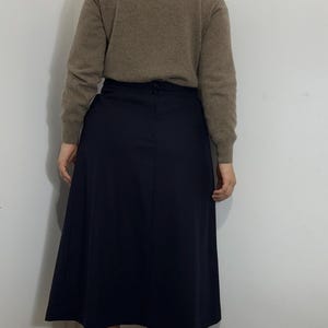 May include: A beige long-sleeved turtleneck sweater paired with a solid navy blue midi skirt. The sweater features a ribbed collar and cuffs, while the skirt has a straight waistband with a visible button closure at the center back.