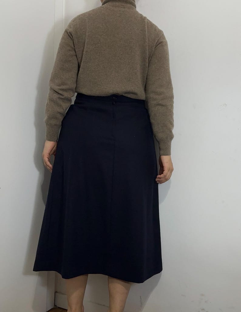 May include: A beige long-sleeved turtleneck sweater paired with a solid navy blue midi skirt. The sweater features a ribbed collar and cuffs, while the skirt has a straight waistband with a visible button closure at the center back.