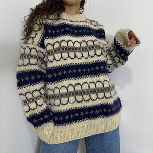 Könnte beinhalten: Cremefarbener Pullover mit einem marineblauen und grauen geometrischen Muster. Der Pullover hat einen Rundhalsausschnitt und lange Ärmel mit gerippten Bündchen und Saum. Das Muster umfasst horizontale Streifen und sich wiederholende Bogenformen.