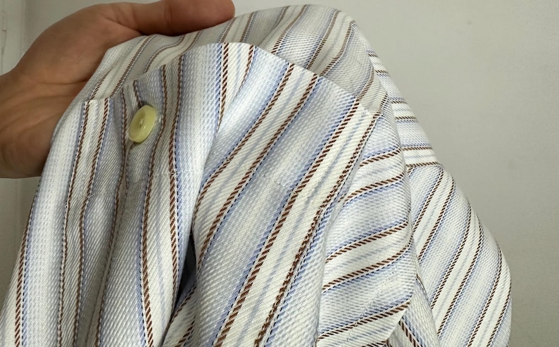May include: A light blue and white striped shirt with brown accent stripes. The shirt has a buttoned cuff and a light green button. The fabric is folded, showing the texture and pattern of the material.