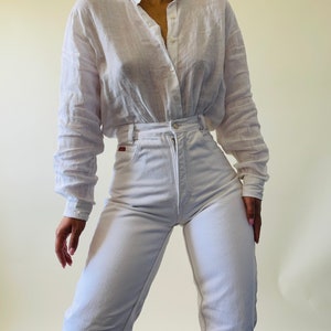 May include: A white button-down shirt and high-waisted white jeans. The shirt has long sleeves and a relaxed fit. The jeans have a classic five-pocket design. The outfit is made of a lightweight fabric.