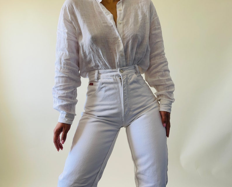 May include: A white button-down shirt and high-waisted white jeans. The shirt has long sleeves and a relaxed fit. The jeans have a classic five-pocket design. The outfit is made of a lightweight fabric.